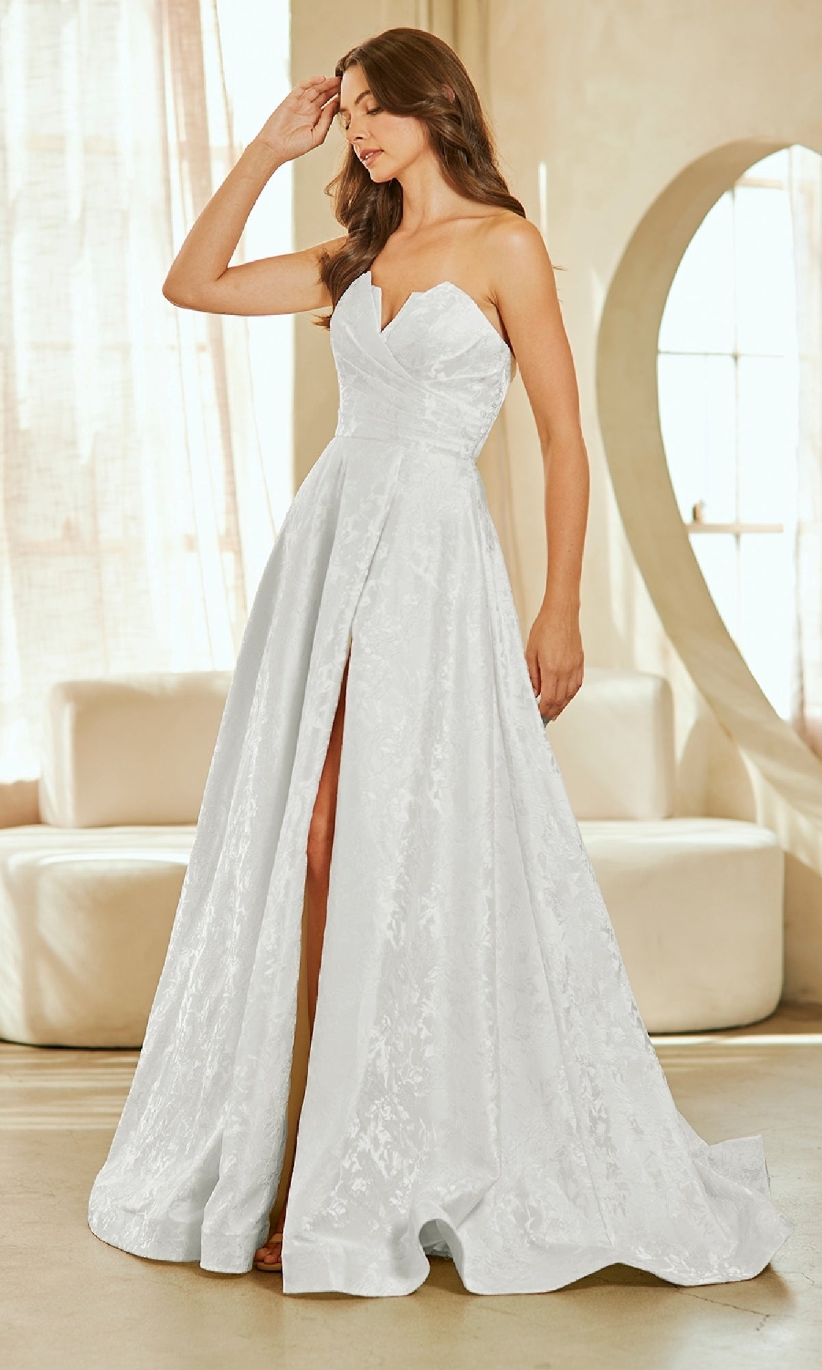 A woman wears the Amelia Couture 8061 Textured Strapless Prom Gown with a sweetheart neckline and front slit, standing indoors near a round mirror and sofa.